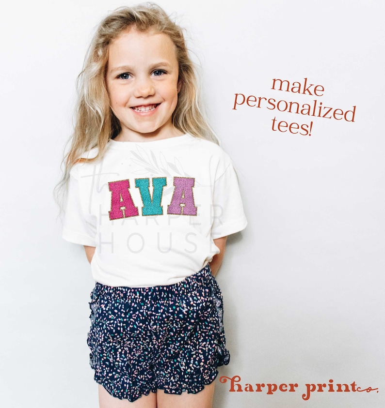 May include: A young girl wearing a white t-shirt with the name "AVA" in colorful letters. The shirt is personalized with a gold glitter outline. The girl is wearing blue and white polka dot shorts.