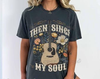 May include: A charcoal gray short-sleeve t-shirt featuring a central acoustic guitar graphic surrounded by various flowers and foliage. The text "THEN SINGS MY SOUL" is printed in a distressed white font above and below the guitar illustration.