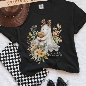 May include: Black t-shirt featuring a cartoon ghost holding a book, surrounded by floral elements. The ghost wears a leopard print bow. Text on the shirt reads "armed & dangerous". A brown cowboy hat and checkered shorts are also in the image.