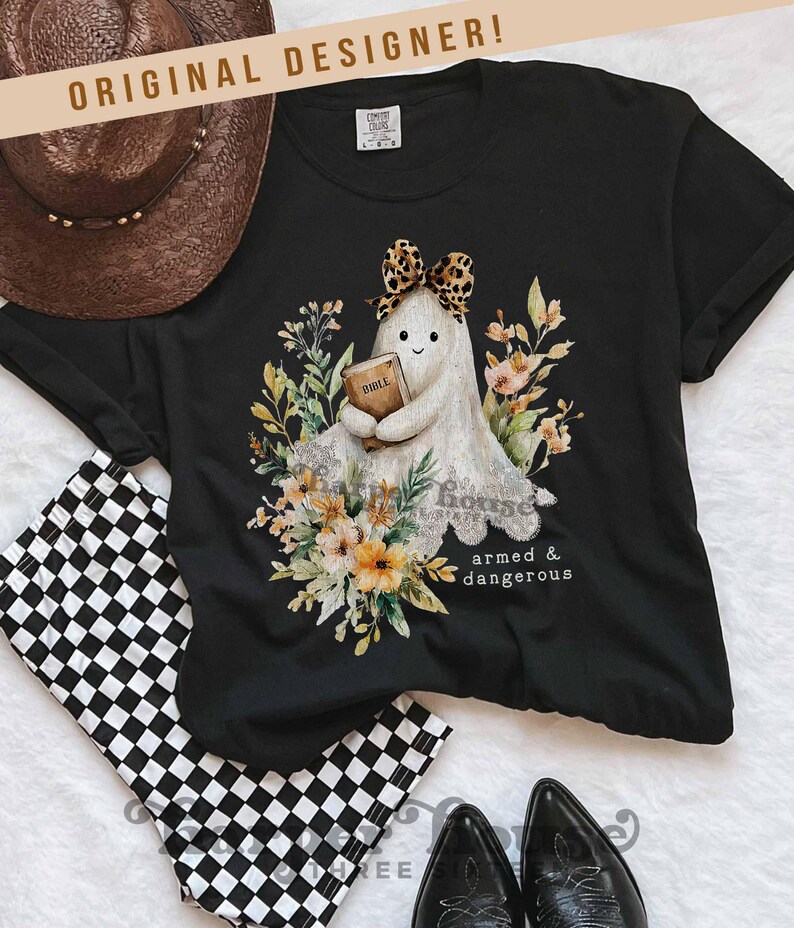 May include: Black t-shirt featuring a cartoon ghost holding a book, surrounded by floral elements. The ghost wears a leopard print bow. Text on the shirt reads "armed & dangerous". A brown cowboy hat and checkered shorts are also in the image.