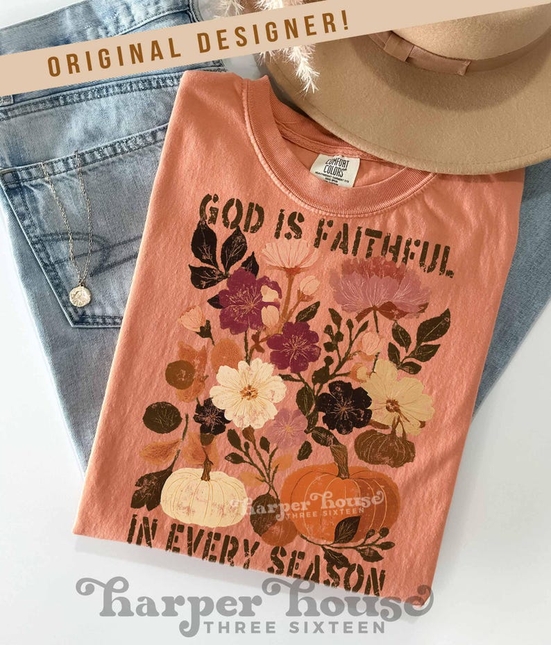 May include: Peach-colored t-shirt with the text "GOD IS FAITHFUL IN EVERY SEASON" and a floral and pumpkin design. The shirt is folded on top of blue jeans, a necklace, and a tan hat. The brand name "Harper House" is also visible.
