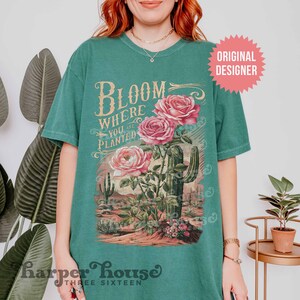 May include: A teal green t-shirt with a vintage-style graphic featuring a cactus and roses. The text on the shirt reads "Bloom Where You Are Planted".