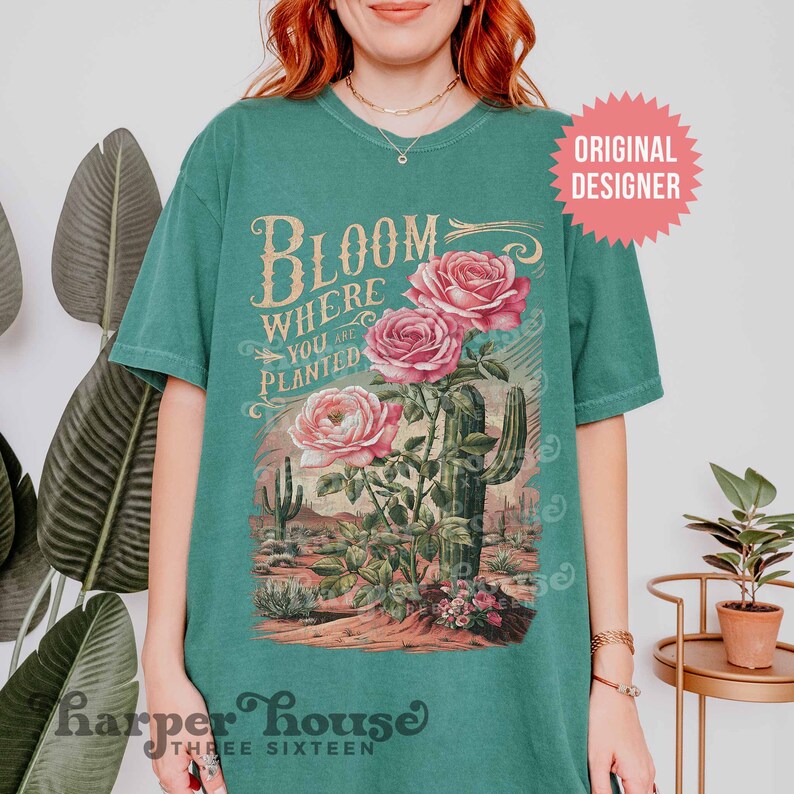 May include: A teal green t-shirt with a vintage-style graphic featuring a cactus and roses. The text on the shirt reads "Bloom Where You Are Planted".