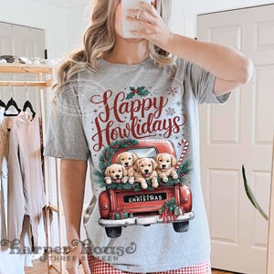 Op de afbeelding: Een grijs T-shirt met de tekst "Happy Howlidays" in rode letters. Op het shirt is een rode vrachtwagen met een kerstkrans en vier golden retriever-puppies gedrukt.