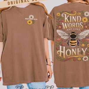 Peut inclure: T-shirt marron avec le texte "Kind Words Are Like Honey" et un graphisme d'abeille au dos. Le devant pr&eacute;sente un petit motif floral et le texte "Proverbes 16:24". Le t-shirt porte &eacute;galement le texte "Harper House Three Sixteen".