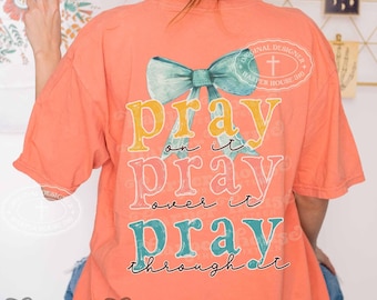 May include: A coral-colored short-sleeve t-shirt featuring a large teal bow graphic and the text "pray on it, PRAY over it, PRAY through it" in yellow, pink, and teal lettering, with a "Harper House 310" logo printed on the back.