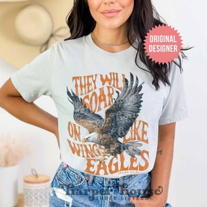 May include: A light blue t-shirt with a vintage-style graphic of an eagle soaring with the text "They will soar on wings like eagles Isaiah 40:31".