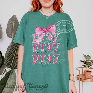 May include: A teal green t-shirt with a pink bow and the text "pray on it pray over it pray through it". The shirt also has a small white cross and the text "Original Designer Harper House 316" in a circle.