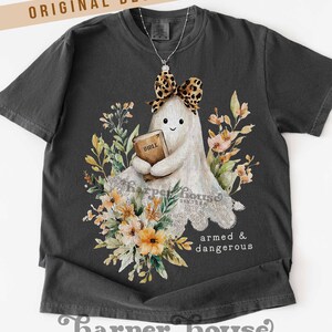 May include: Dark gray t-shirt featuring a cartoon ghost holding a Bible, adorned with a leopard print bow and surrounded by floral elements. The text "armed & dangerous" is printed on the shirt. The brand name "Harper House Three Sixteen" is at the bottom.
