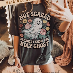 May include: Dark gray t-shirt with the text "NOT SCARED BECAUSE I KNOW THE HOLY GHOST" and a cartoon ghost illustration surrounded by flowers. The shirt has short sleeves and a relaxed fit.