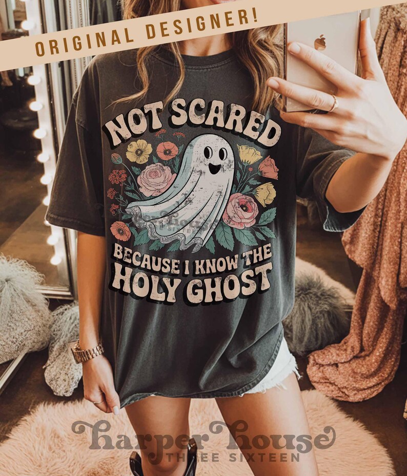 May include: Dark gray t-shirt with the text "NOT SCARED BECAUSE I KNOW THE HOLY GHOST" and a cartoon ghost illustration surrounded by flowers. The shirt has short sleeves and a relaxed fit.