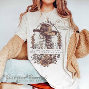 May include: A cream-colored short-sleeve t-shirt featuring a brown graphic of a person wearing a cowboy hat with a braided hairstyle. The design includes floral elements and the text: "perhaps you were born for SUCH A TIME AS THIS" and "Esther 4:14".