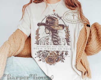 May include: A cream-coloured short-sleeve t-shirt featuring a brown graphic of a person wearing a cowboy hat with a braided hairstyle. The design includes floral elements and the text: "perhaps you were born for SUCH A TIME AS THIS" and "Esther 4:14".