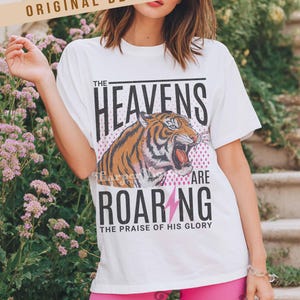 May include: White t-shirt with a tiger graphic and the text "THE HEAVENS ARE ROARING". The tiger is orange and black with a roaring mouth. The text "THE PRAISE OF HIS GLORY" is below the text "ROARING".