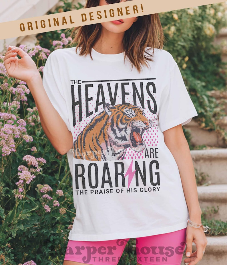 May include: White t-shirt with a tiger graphic and the text "THE HEAVENS ARE ROARING". The tiger is orange and black with a roaring mouth. The text "THE PRAISE OF HIS GLORY" is below the text "ROARING".