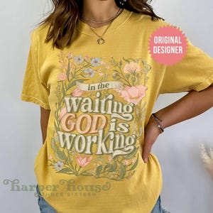 May include: Yellow t-shirt with a floral design and the text "In the waiting God is working". The shirt has a distressed look and is made by Harper House Three Sixteen.