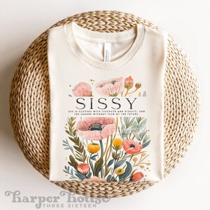 May include: A cream-colored t-shirt with a floral design and the text "Sissy" in black. The text below reads "She is clothed with strength and dignity, and she laughs without fear of the future."