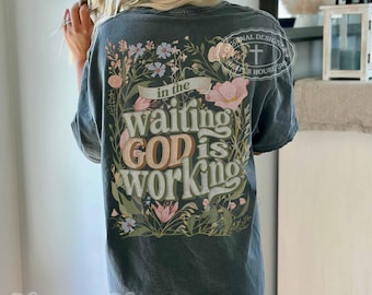Puede incluir: Una camiseta gris carbón de manga corta con un gráfico floral en la espalda con el texto "in the waiting GOD is working" y un pequeño logotipo de cruz. El diseño incluye varias flores silvestres en tonos rosa, azul y verde.