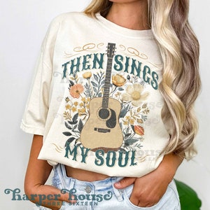 May include: A white t-shirt with a floral design and an acoustic guitar. The text "Then Sings My Soul" is printed on the shirt.