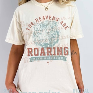 May include: A cream-colored short-sleeve t-shirt featuring a graphic of a tiger head in the center. The text reads THE HEAVENS ARE ROARING and THE PRAISE OF HIS GLORY, accompanied by a small cross symbol below the tiger illustration.