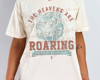 May include: A cream-coloured short-sleeve t-shirt featuring a graphic of a tiger head in the centre. The text reads THE HEAVENS ARE ROARING and THE PRAISE OF HIS GLORY, accompanied by a small cross symbol below the tiger illustration.