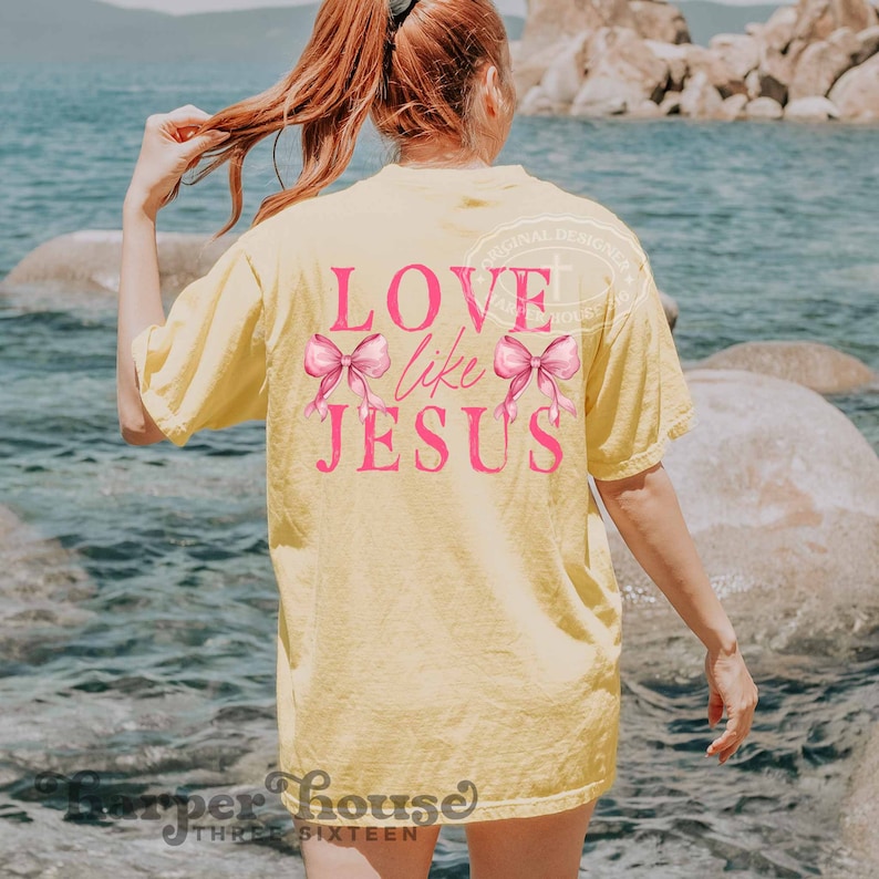 K&ouml;nnte beinhalten: Ein hellgelbes T-Shirt mit dem Text "LOVE like JESUS" in Pink, verziert mit zwei rosa Schleifen. Das Shirt hat eine lockere Passform. Der Hintergrund zeigt Wasser und Felsen.