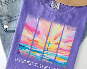 May include: A purple short-sleeve t-shirt featuring a colorful abstract sunset graphic with a central cross. The text "WASHED IN THE WATER" is printed in white below the graphic. The shirt has a Comfort Colors label visible at the neck.
