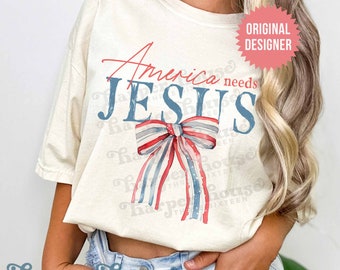Op de afbeelding: Een wit T-shirt met de tekst "America needs JESUS" in blauwe en rode letters. Een rood-wit-blauwe strik is gecentreerd op het shirt.