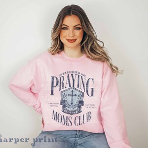 Op de afbeelding: Roze trui met een blauwe afbeelding waarop staat "Established 2003 Praying Moms Club" met een kruis en een krans. De afbeelding heeft de tekst "A Mother's Prayers Redeemed Can Shake The World."