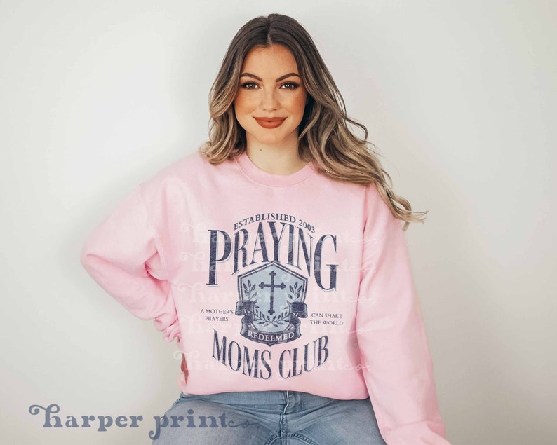 Op de afbeelding: Roze trui met een blauwe afbeelding waarop staat "Established 2003 Praying Moms Club" met een kruis en een krans. De afbeelding heeft de tekst "A Mother's Prayers Redeemed Can Shake The World."