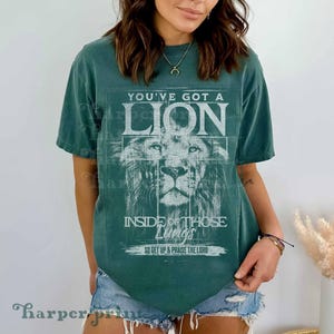 May include: A dark teal short-sleeve t-shirt featuring a white graphic of a lion's face. The text reads: YOU'VE GOT A LION INSIDE OF THOSE LUNGS SO GET UP & PRAISE THE LORD. The shirt has a crew neckline and a relaxed fit.