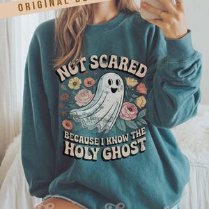 May include: Teal-colored sweatshirt with the text "NOT SCARED BECAUSE I KNOW THE HOLY GHOST" and a cartoon ghost illustration. The design includes floral elements in various colors. The brand name "Harper House Three Sixteen" is at the bottom.