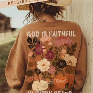 May include: A brown sweatshirt with floral and pumpkin graphics. The back of the sweatshirt features the text "GOD IS FAITHFUL IN EVERY SEASON". The design includes pink, white, and burgundy flowers, and orange and white pumpkins.