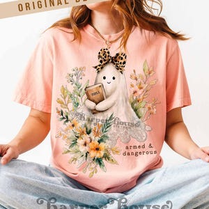 May include: Peach-colored t-shirt featuring a cartoon ghost holding a book labeled "Bible" and wearing a leopard print bow. The shirt has floral accents and the text "armed & dangerous". The brand name "Harper House" is also visible.
