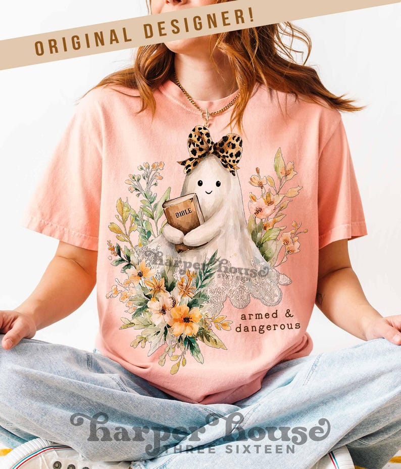 May include: Peach-colored t-shirt featuring a cartoon ghost holding a book labeled "Bible" and wearing a leopard print bow. The shirt has floral accents and the text "armed & dangerous". The brand name "Harper House" is also visible.
