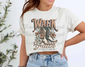May include: A cream-colored short-sleeve t-shirt featuring a graphic print of two cowboy boots surrounded by flowers. The text reads WALK BY FAITH Even When You Cannot See 2 Corinthians 5:7 in brown and orange lettering.
