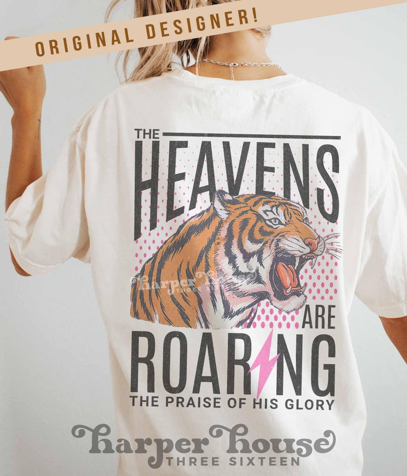 May include: Cream-colored t-shirt with a tiger graphic. The design includes the words "THE HEAVENS ARE ROARING" and "THE PRAISE OF HIS GLORY". The tiger is orange and black with its mouth open. The shirt also has the text "ORIGINAL DESIGNER!" at the top.