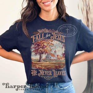 May include: Navy blue t-shirt with a fall-themed graphic. The graphic features a church, a tree with autumn leaves, and the text "Fall for Jesus" and "He Never Leaves". The shirt also has the text "Harper House Three Sixteen".