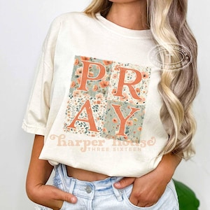 Op de afbeelding: Een crèmekleurig t-shirt met korte mouwen en een grote vierkante afbeelding met het woord PRAY in dikke oranje letters. De letters liggen op een achtergrond van kleine oranje en groene bloemmotieven. De tekst Harper House THREE SIXTEEN staat onder de afbeelding.