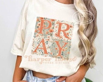Puede incluir: Una camiseta de manga corta color crema con un gran gráfico cuadrado que muestra la palabra PRAY en letras naranjas en negrita. Las letras están superpuestas sobre un fondo de pequeños patrones florales naranjas y verdes. El texto Harper House THREE SIXTEEN aparece impreso debajo del gráfico.