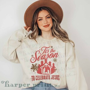 May include: A cream colored sweatshirt with the text "'Tis the Season to Celebrate Jesus" in red and gold lettering. The design includes a red silhouette of a manger scene with a gold forest behind it.