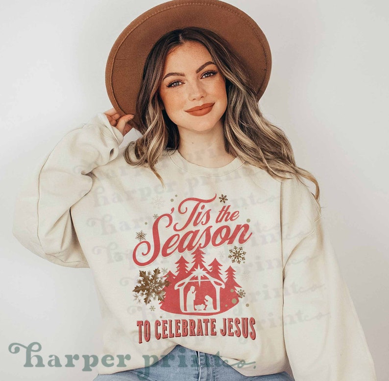May include: A cream colored sweatshirt with the text "'Tis the Season to Celebrate Jesus" in red and gold lettering. The design includes a red silhouette of a manger scene with a gold forest behind it.