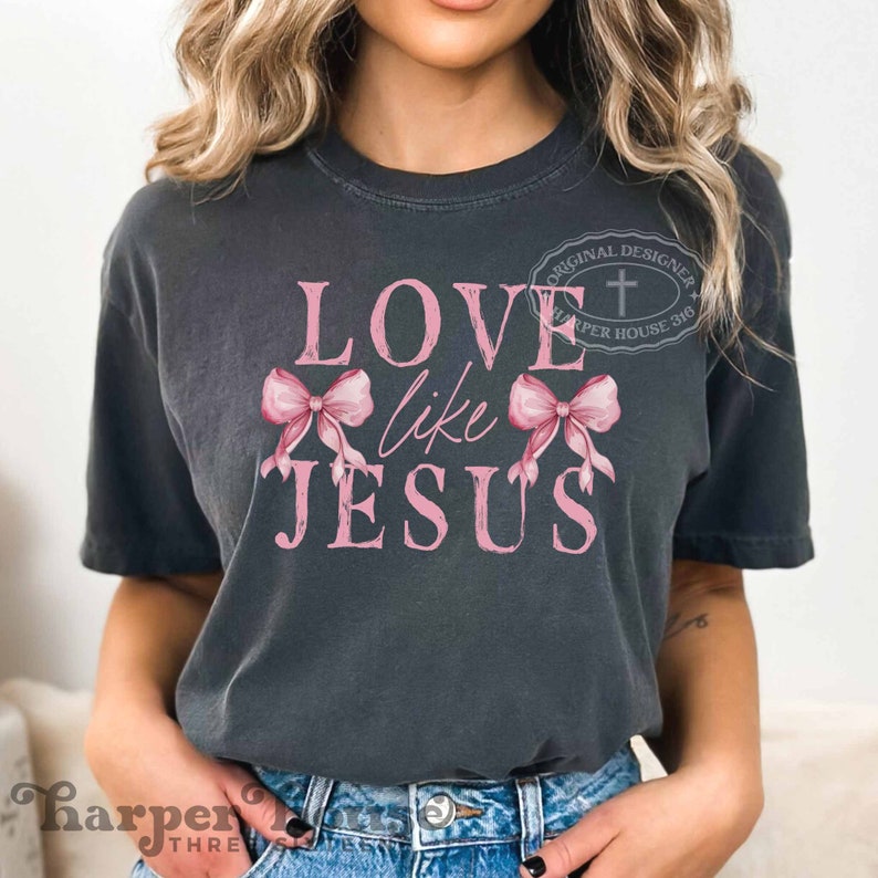 K&ouml;nnte beinhalten: Dunkelgraues T-Shirt mit dem rosa Text "LOVE like JESUS" und rosa Schleifengrafiken. Das Shirt hat einen Vintage-Look, und das Design enth&auml;lt ein Kreuzlogo und den Text "ORIGINAL DESIGNER HARPER HOUSE 316".