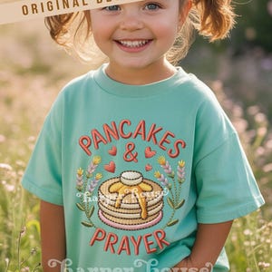 May include: A light teal t-shirt with the embroidered words "PANCAKES & PRAYER" in red script. The design includes a stack of pancakes with syrup, hearts, and floral accents. The brand name "Harper House" is at the bottom.