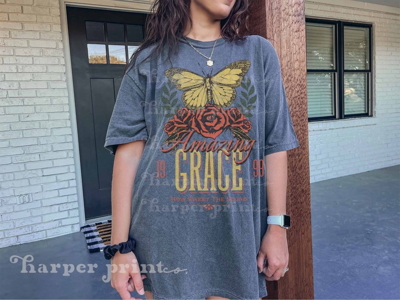Pode incluir: Uma camiseta cinza com uma estampa gr&aacute;fica de uma borboleta, flores e o texto "Amazing Grace 99 How Sweet The Sound".