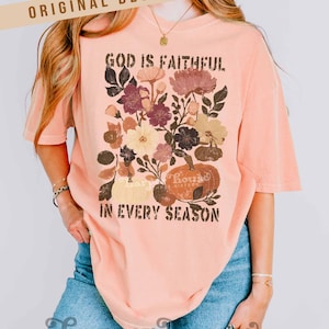 May include: Peach-colored t-shirt with a floral and pumpkin graphic. The design includes the text "GOD IS FAITHFUL IN EVERY SEASON." The shirt has short sleeves and a relaxed fit. The words "ORIGINAL DESIGNER!" are at the top.