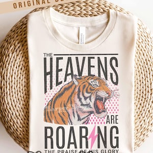 May include: Cream-colored t-shirt with a graphic of a roaring tiger. The text reads "THE HEAVENS ARE ROARING" with a pink lightning bolt and the words "THE PRAISE OF HIS GLORY". The shirt has a crew neck and is made of a soft material.