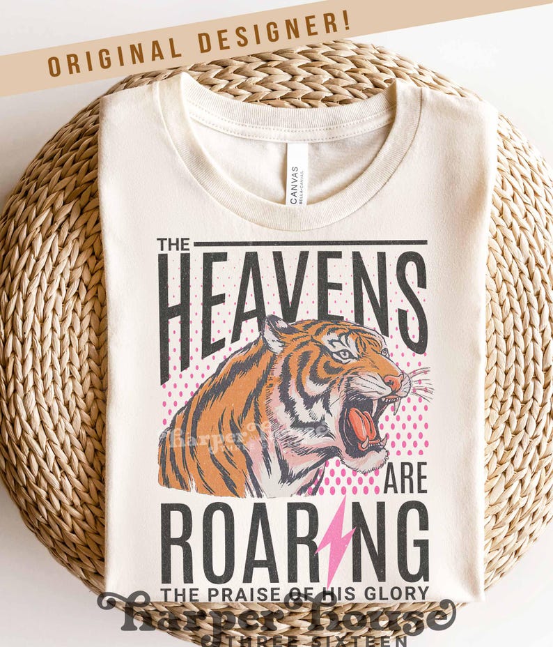 May include: Cream-colored t-shirt with a graphic of a roaring tiger. The text reads "THE HEAVENS ARE ROARING" with a pink lightning bolt and the words "THE PRAISE OF HIS GLORY". The shirt has a crew neck and is made of a soft material.