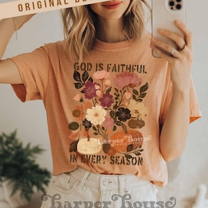 May include: Peach-colored t-shirt with a floral and pumpkin graphic. The text on the shirt reads "GOD IS FAITHFUL IN EVERY SEASON." The design includes various flowers and pumpkins. The words "ORIGINAL DESIGNER!" are at the top.