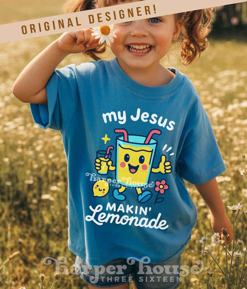 Op de afbeelding: Een blauw t-shirt met de tekst "my Jesus makin' lemonade" en een cartoon limonade afbeelding. Het shirt wordt gedragen door een kind dat een madeliefje vasthoudt. De woorden "Original Designer" staan bovenaan.